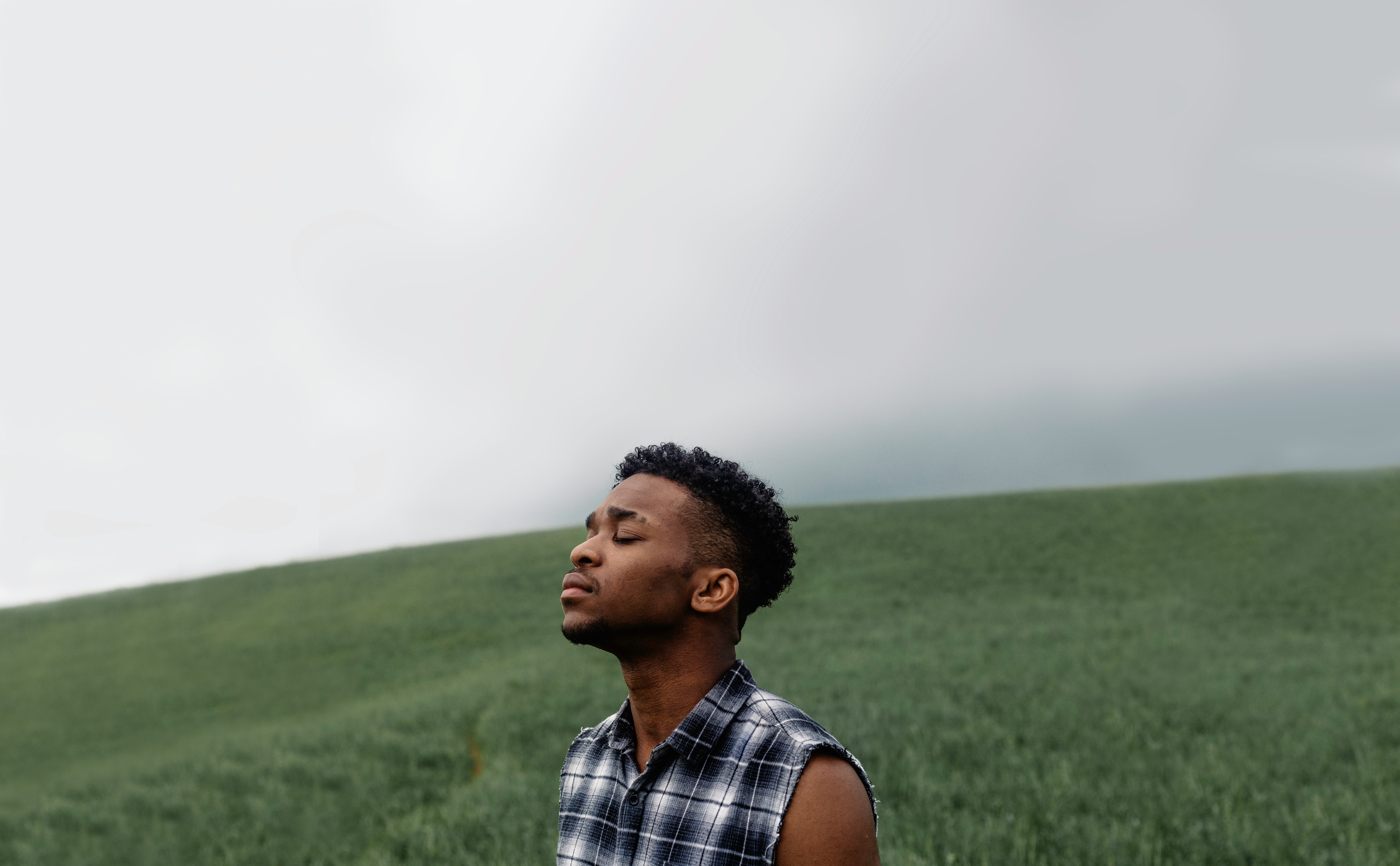 A dark skinned, masc presenting individual with a plaid tank top and pained expression tilts their face up, eyes closed, toward a grey sky. They are standing in a grassy field. We see only their shoulders and up.