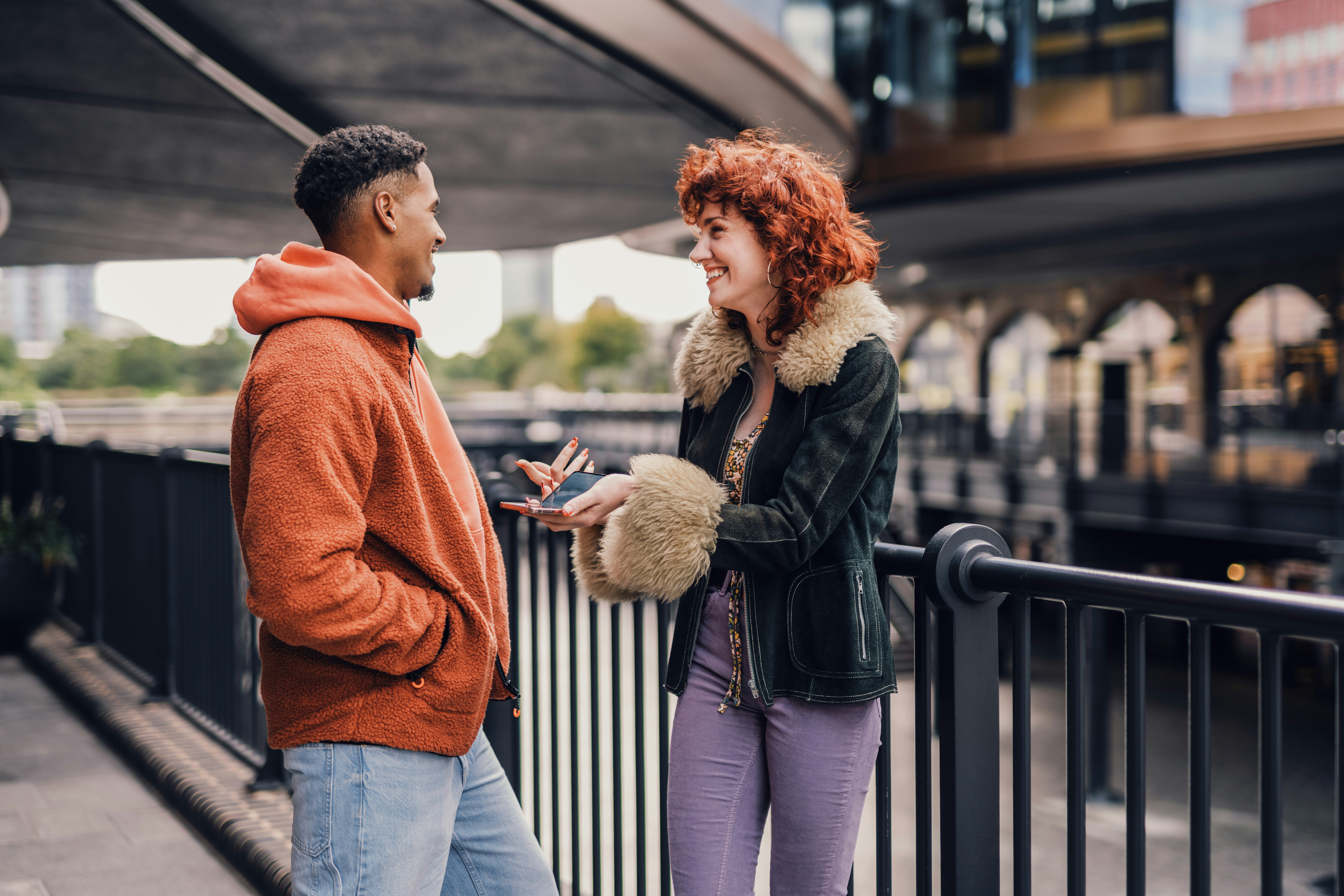 Stock photo of two young people chatting in an urban setting with a city out-of-focus on the background. One is a masc Black person and the other is a femme white person who holds their phone in their hand as they gesticulate. Both are smiling.